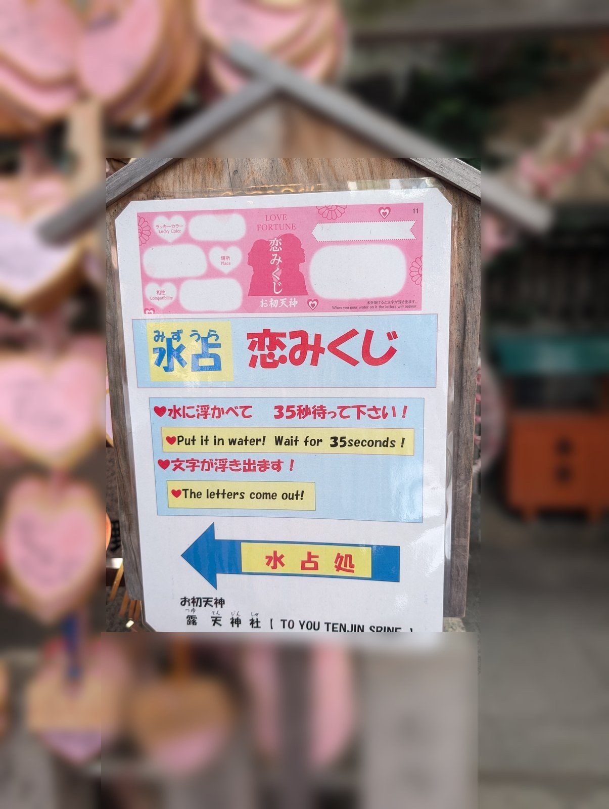 2026年8月6日撮影。境内の案内看板です。恋みくじの見本の下に「水に浮かべて35秒待って下さい」「文字が浮き出ます」と書かれ、水占処への矢印が出ています。