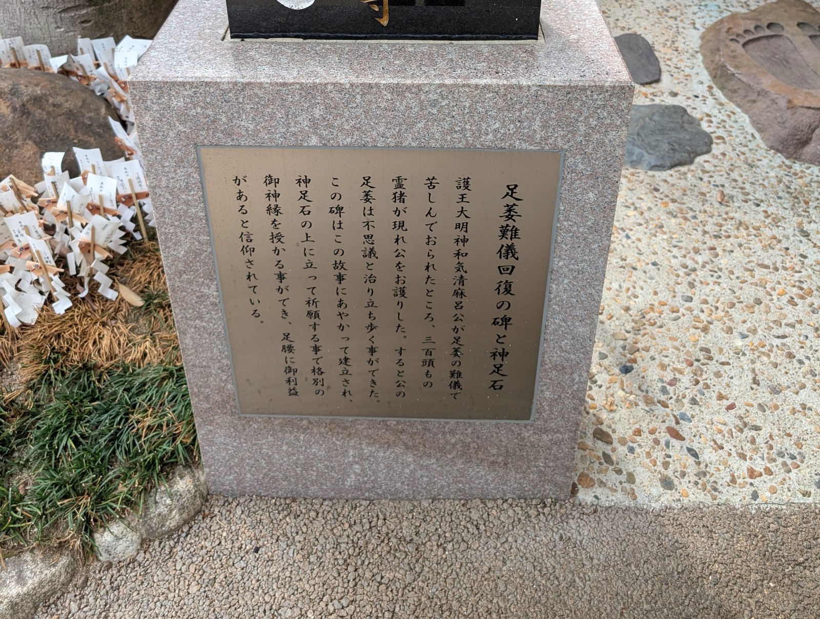 現地の説明板「足萎難儀回復の碑と神足石」。右奥に足形の刻まれた石(2026-08-22 撮影)